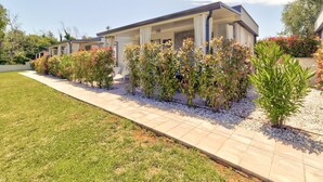 A modern residential building with a covered patio area, a well-maintained lawn, and various flowering shrubs. - Mobile Home in Banjole near Beach & Pool (Banjole)