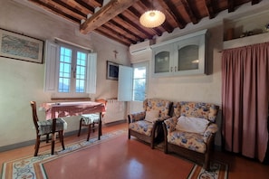 A cozy room with a wooden ceiling, a floral sofa, a dining table with checkered tablecloth, and a window with curtains.