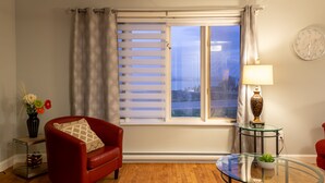 A living room with a red armchair, a glass coffee table, a vase with flowers, and a window with blinds. - Maison des Champs (Saint-Jean-de-l'Île-d'Orléans)
