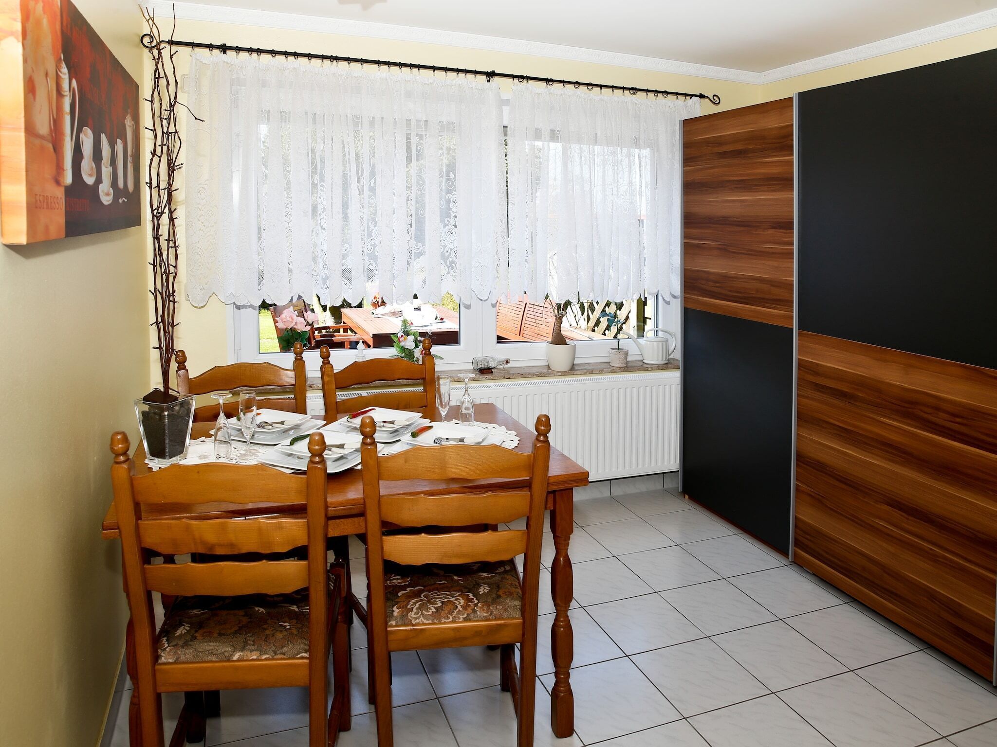 A dining area with a wooden table set for four, a window with lace curtains, and a view of a garden.