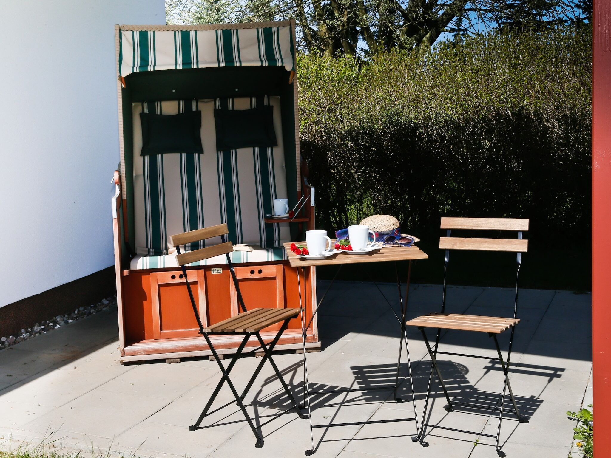An outdoor seating area with a striped awning, two wooden chairs, a table with a cup and a plate of strawberries, and a bench.
