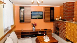A living room with a sofa, a coffee table with a book and a bottle, a TV mounted on the wall, a fireplace, and a wooden cabinet. - Cozy Baltic Escape for 2 Guests (Bastorf / Kägsdorf)