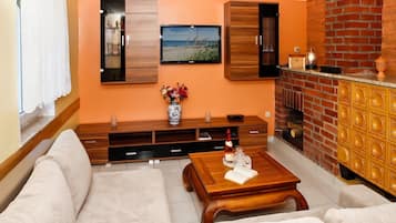 Een woonkamer met een bank, een bijzettafeltje met een boek en een fles, een aan de muur bevestigde televisie, een open haard en een houten kast.