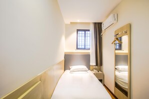 Habitació d'hotel estreta amb un llit individual, una taula de nit amb un telèfon, una finestra amb cortines i un aire condicionat fixat a la paret.