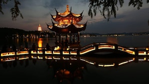 Un pabellón chino tradicional junto a un lago tranquilo al anochecer, con una pagoda distante y un puente que refleja las luces.