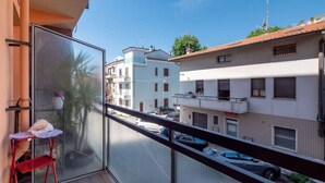 Un balcon avec une chaise rouge et une petite table, donnant sur une rue bordée de voitures garées et d’immeubles.