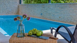 A poolside setting with a wicker table holding a vase with flowers, grapes, and apples.