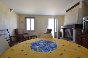 A dining area with a round table covered by a yellow tablecloth with blue floral patterns, flanked by wooden chairs, a small table with a glass, and a fireplace.