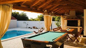 Una mesa de billar con superficie de fieltro verde, sillas de madera y una mesa con cubierta blanca. Hay una piscina con agua azul cristalina y un techo de madera con lámparas colgantes.