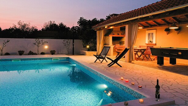 Una piscina con sillas de descanso y mesa de billar, rodeada por un patio con techo de baldosas.