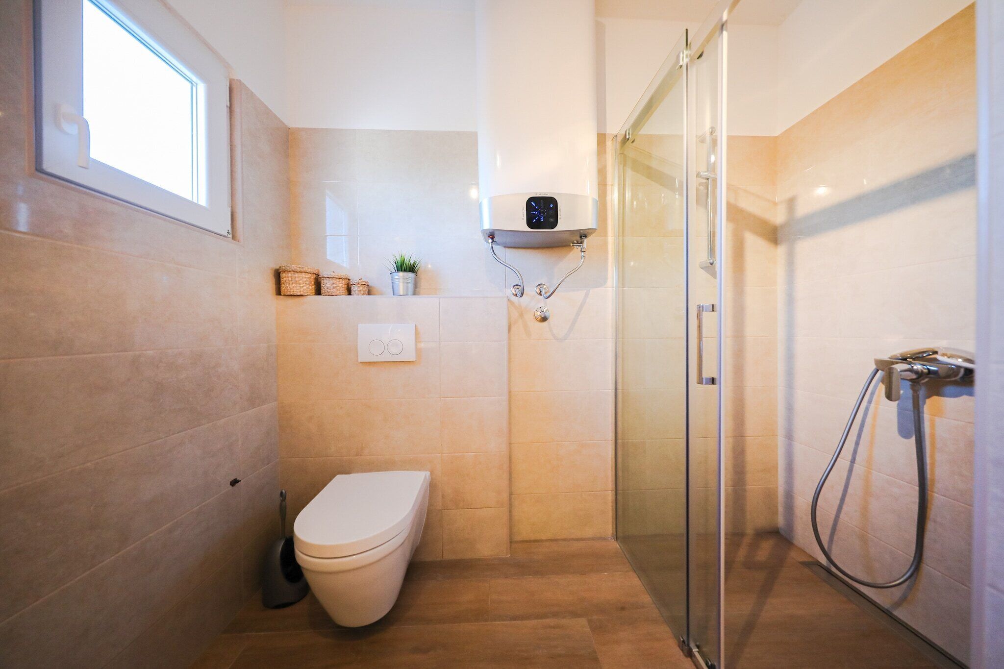 A modern bathroom with a toilet, a shower, and a glass-enclosed shower stall. There is a shelf with a small plant and a power outlet.