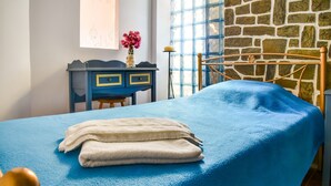 A bedroom with a blue bedspread, a wooden headboard, a desk with drawers, a vase with flowers, and a stone wall. - Alluring Holiday Home in Syros with Barbecue (Syros)