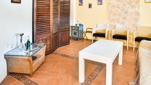A living room with a glass-top table, wicker sofa, white coffee table, and a stone wall. There are two chairs and a small table with a bottle and glasses. - Alluring Holiday Home in Syros with Barbecue (Syros)
