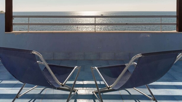A balcony with two blue chairs facing a calm sea under a clear sky.