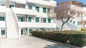 A multi-story building with a staircase leading to the entrance, a flat roof with a pergola, and a hedge in the foreground.