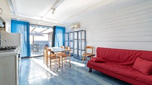 A compact living space with a kitchenette, a dining area with a table and chairs, a red sofa, and a view through a large window.