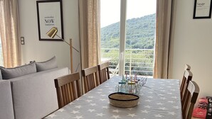 A dining area with a table covered in a star-patterned tablecloth, wooden chairs, and a view of a mountainous landscape through large windows. - Villa with pool, between olive trees and sea - fantastic view! (Cargèse)