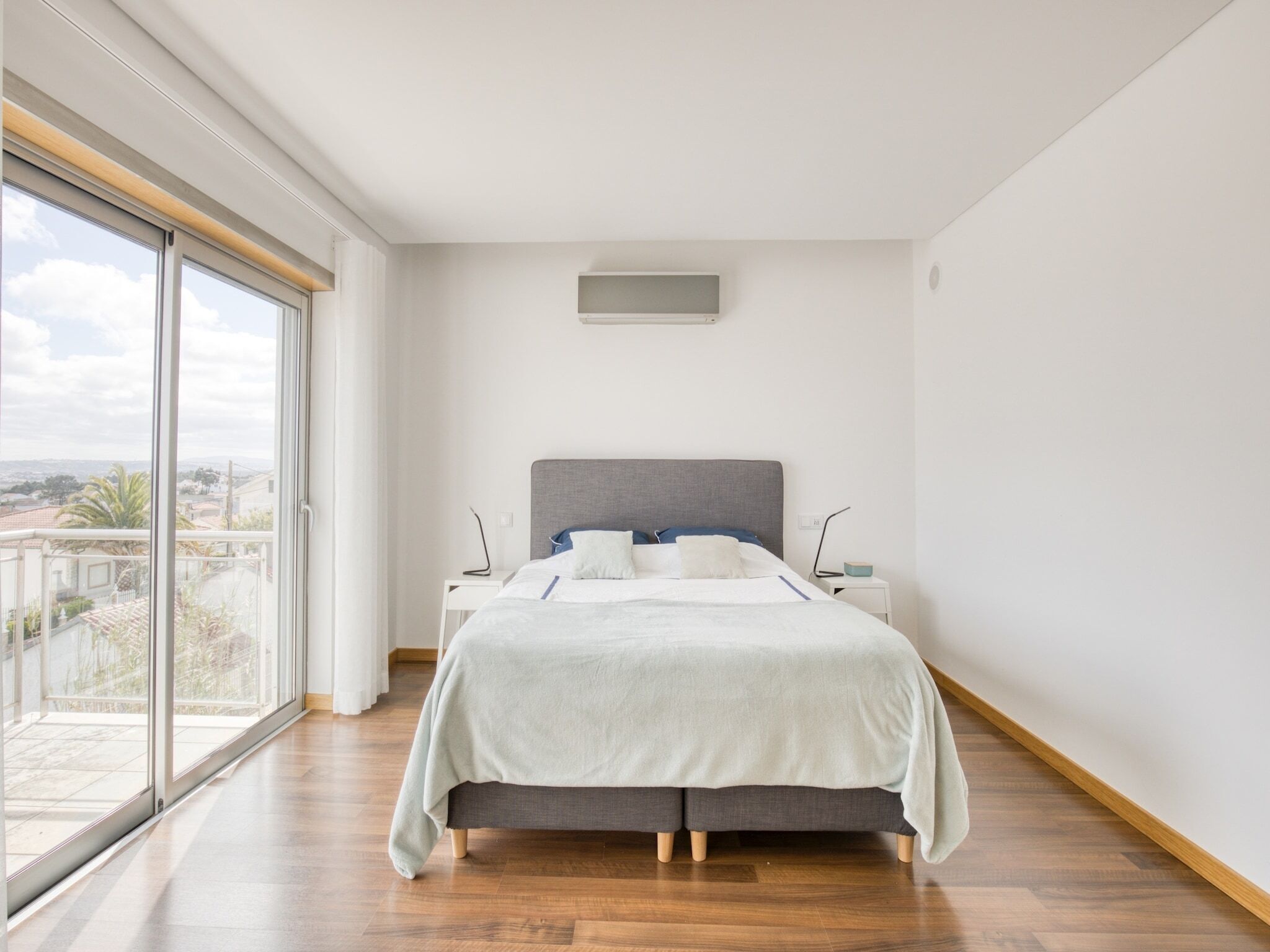 A bedroom with a large window, a bed with a grey headboard, and a view of the city.