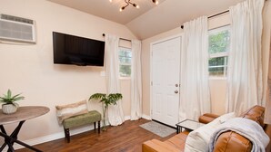 A living room with a brown leather sofa, a green bench, a small table with a plant, a flat-screen TV mounted on the wall, and a white door with curtains. - ⫷1 BD/1 Bath⫷Tiny Magnolia Retreat⫷⫸Near TTU⫸ (Lubbock)