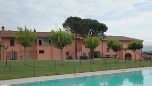 Una piscina con superficie de agua azul cristalina, rodeada por una valla. Al fondo, un edificio con tejado de baldosas y un gran árbol en su fachada.