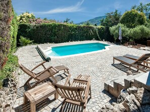 A rectangular swimming pool surrounded by a paved patio with wooden lounge chairs and a table.