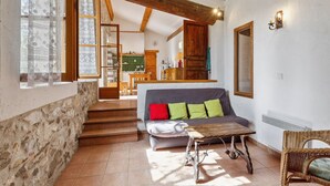 Una sala de estar acogedora con un sofá, una mesa rústica y una silla de mimbre. Una escalera conduce a un nivel superior donde se ve un área de cocina.
