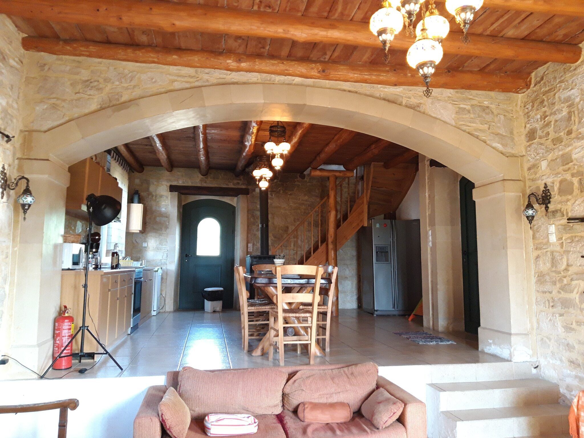 A cozy interior with a stone archway leading to a dining area, wooden beams on the ceiling, and a modern kitchen with a refrigerator.