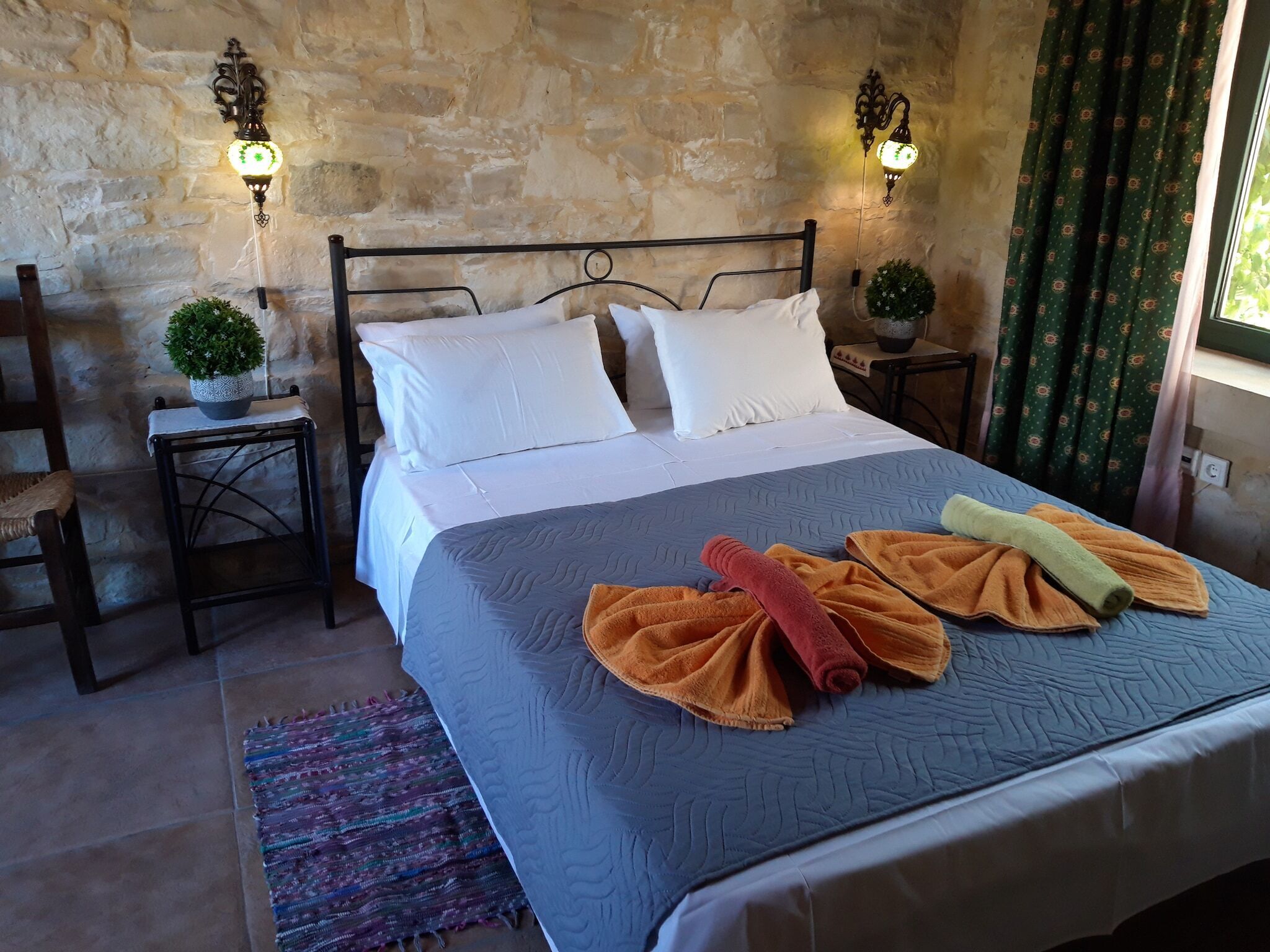 A bed with white pillows, a grey quilt, and rolled towels in orange and yellow. There are two wall-mounted lights, a small potted plant, and a chair with a woven seat.