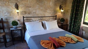 Una cama con almohadas blancas, un edredón gris y toallas enrolladas en naranja y amarillo. Hay dos luces fijadas en la pared, una pequeña planta en maceta y una silla con asiento tejido.