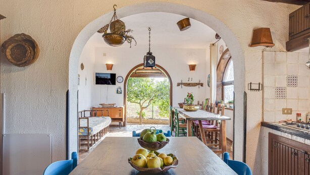 A dining area with a long wooden table, blue chairs, and a bowl of fruit. An arched doorway leads to a kitchen with a tiled backsplash and a hanging light fixture.