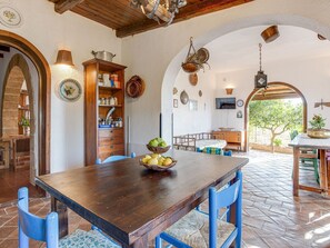 Un coin repas rustique avec une table en bois, des chaises bleues et un saladier rempli de fruits. On y trouve également un buffet en bois avec des bocaux et un téléviseur accroché au mur.