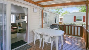 A covered outdoor area with a wooden deck, a white table with chairs, and a sliding glass door leading to an interior space with a couch and a microwave.