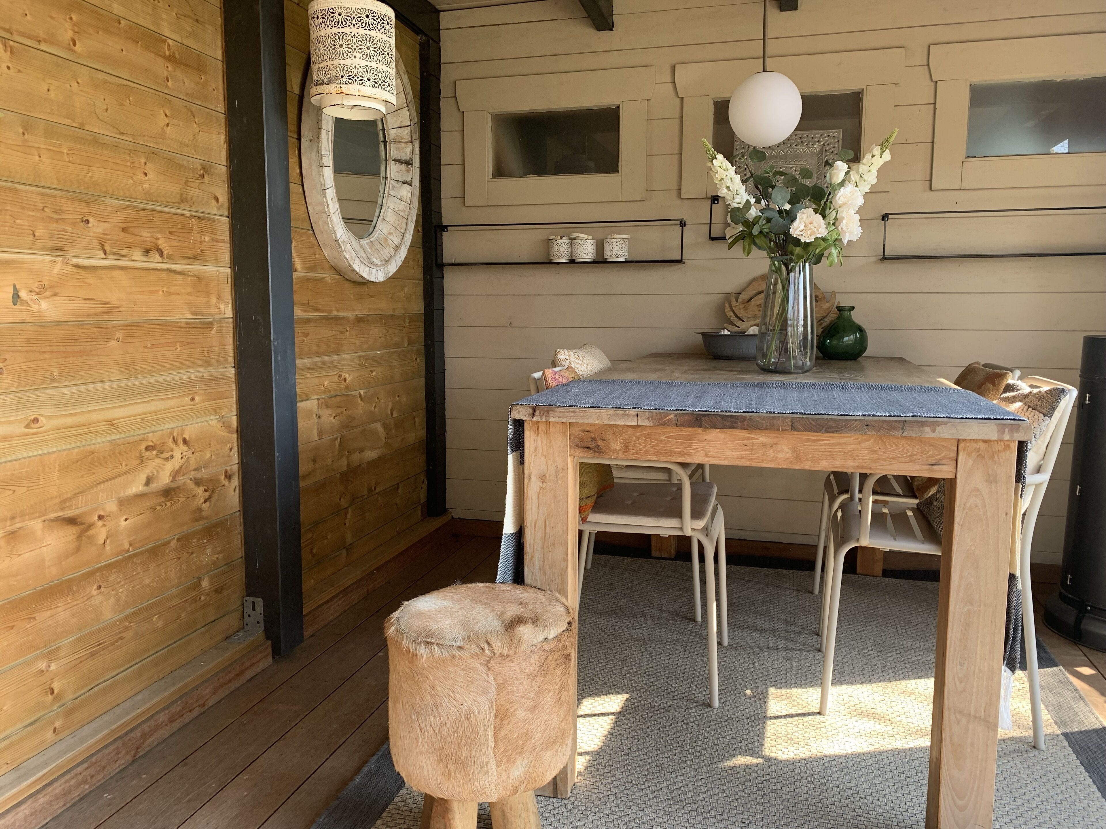 A wooden interior with a round mirror, a table with chairs, a vase with flowers, and a hanging light fixture.