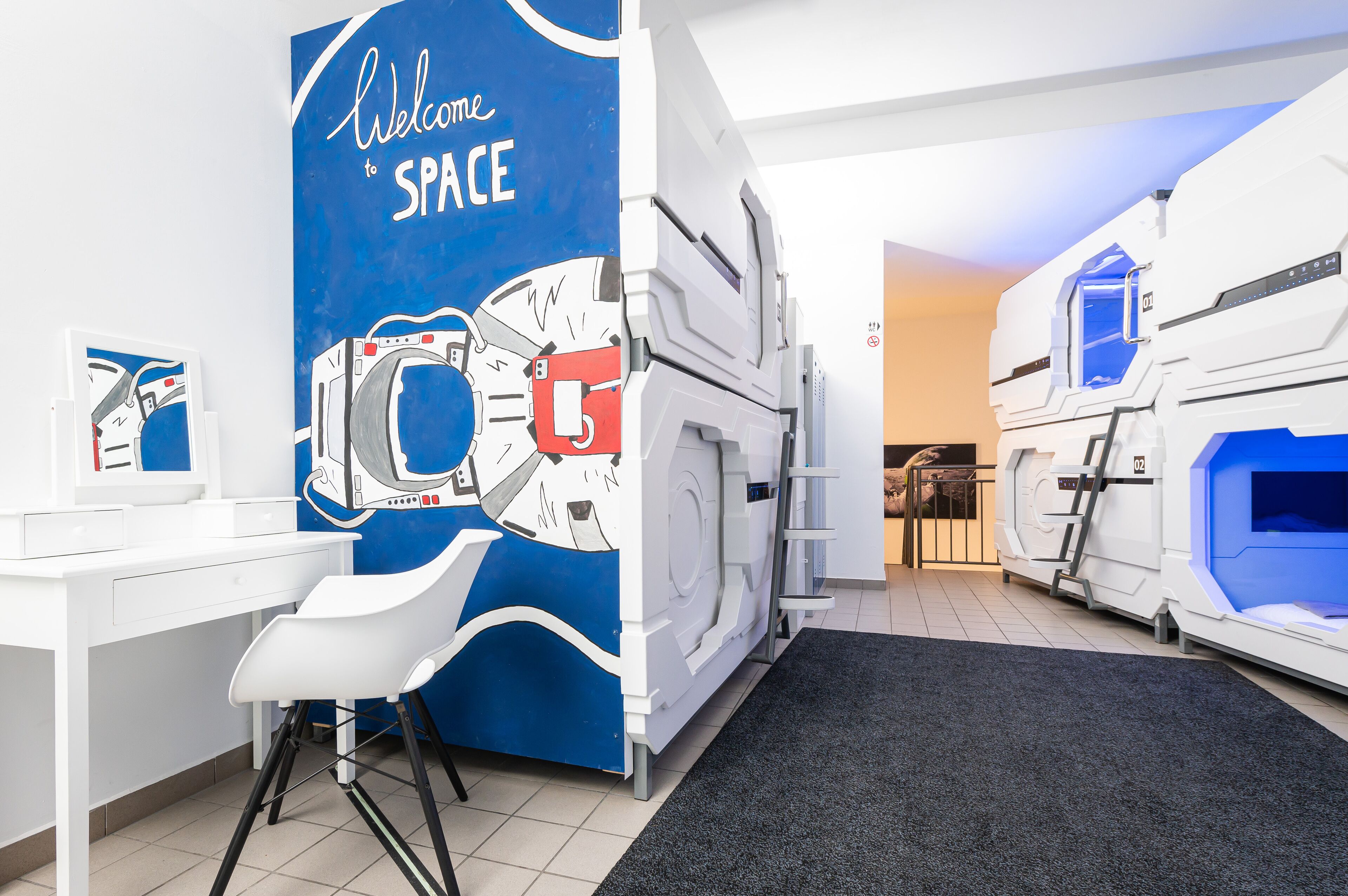A room with a blue wall featuring a space-themed mural, a white desk with a chair, and two large, futuristic-looking beds.