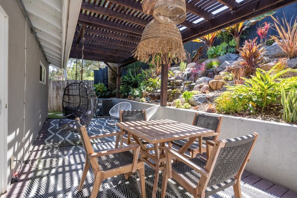 Une terrasse extérieure avec des meubles en bois, un luminaire suspendu et un jardin agrémenté de diverses plantes et de rochers.
