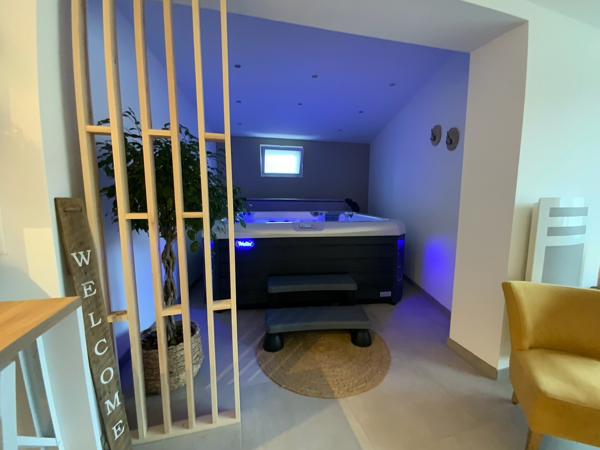 Een moderne kamer met een jacuzzi, een houten scheidingswand met het opschrift 'WELCOME', een potplant en een gele stoel.