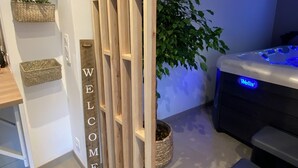 A modern room with a wooden partition labeled "WELCOME," a small plant, a hot tub with a blue light, and a woven basket. - Gîte with private Jacuzzi (St martin choquel)