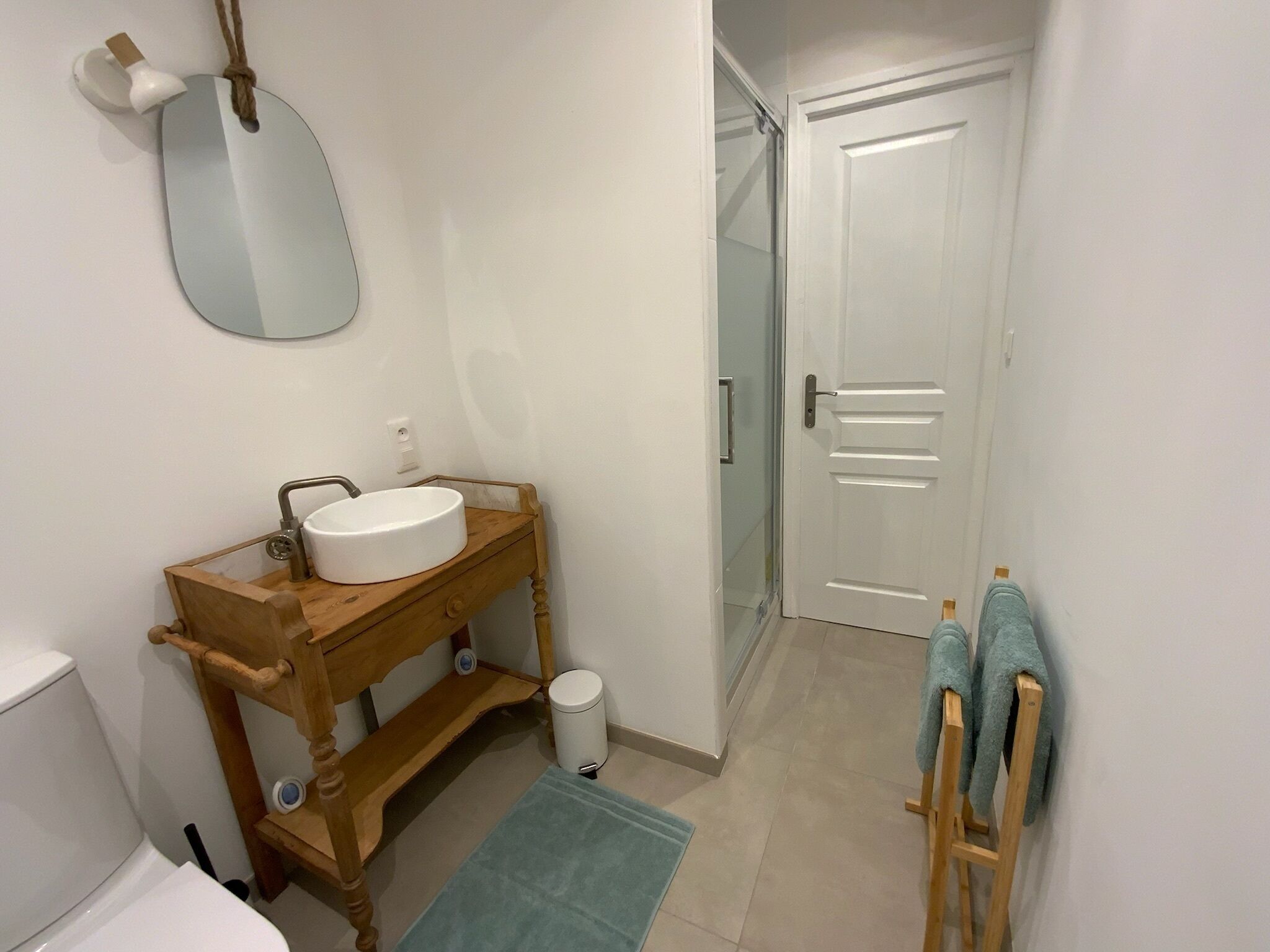 Une salle de bain avec un meuble-lavabo en bois, un lavabo blanc, un miroir, des toilettes, une poubelle et un porte-serviettes.