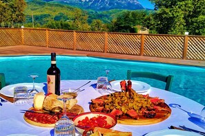 Une table à manger dressée avec une variété de plats et de boissons, placée au bord d’une piscine, avec un paysage montagneux en arrière-plan.