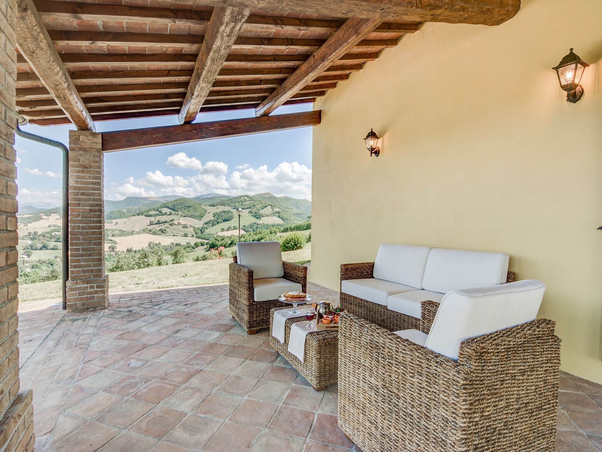 A covered patio with wicker furniture, a table with drinks, and a view of rolling hills.