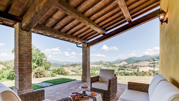 A covered outdoor seating area with wicker furniture and a view of rolling hills. - Holiday Home in Urbania with Hill Views (Urbania)