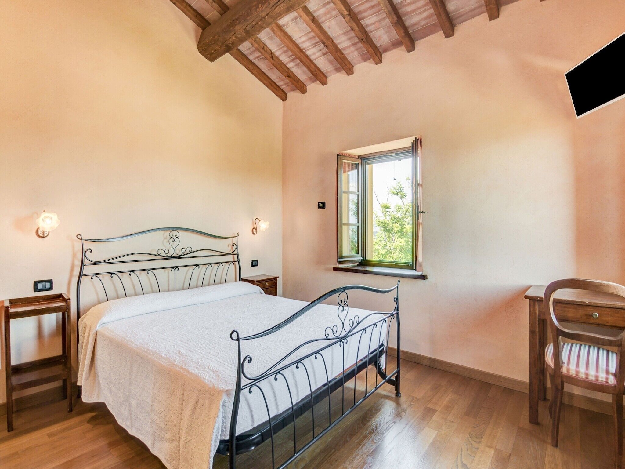 Une chambre à coucher avec un lit en métal, une chaise en bois, une table de chevet et une commode, ainsi qu’une fenêtre donnant sur des arbres.