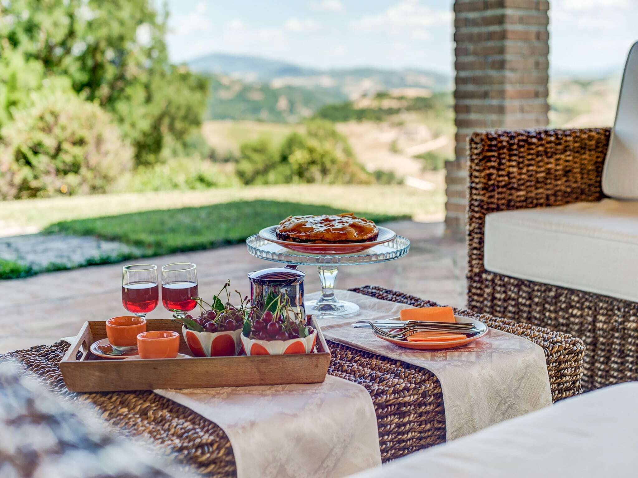 Un espace de restauration en plein air avec une chaise en osier, une table sur laquelle se trouve un plateau de nourriture, des verres, et une vue panoramique en arrière-plan.