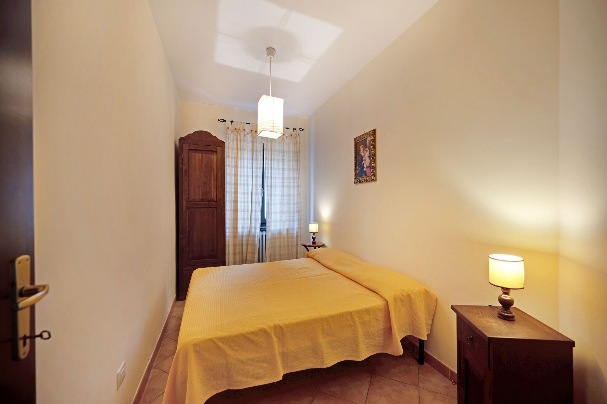 Habitación de hotel estrecha con cama, mesita de noche con lámpara, puerta y ventana con cortinas.