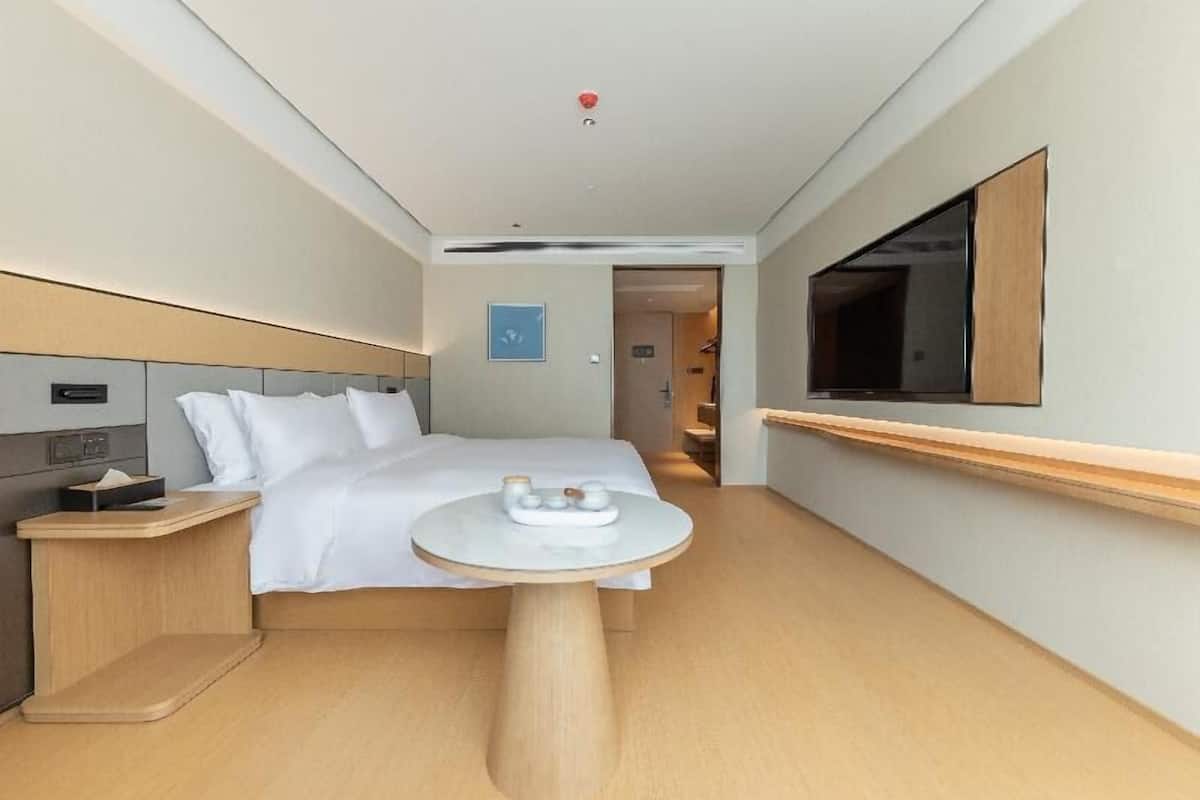 A hotel room with a bed, bedside table, a small round table with a tray, a TV mounted on the wall, and a door leading to another room.