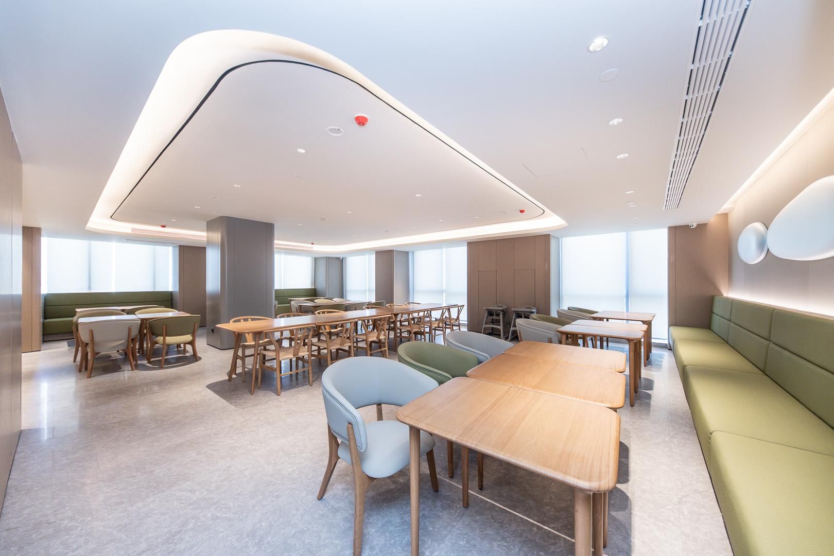 A modern dining area with multiple tables and chairs, a high ceiling with recessed lighting, and large windows providing natural light.