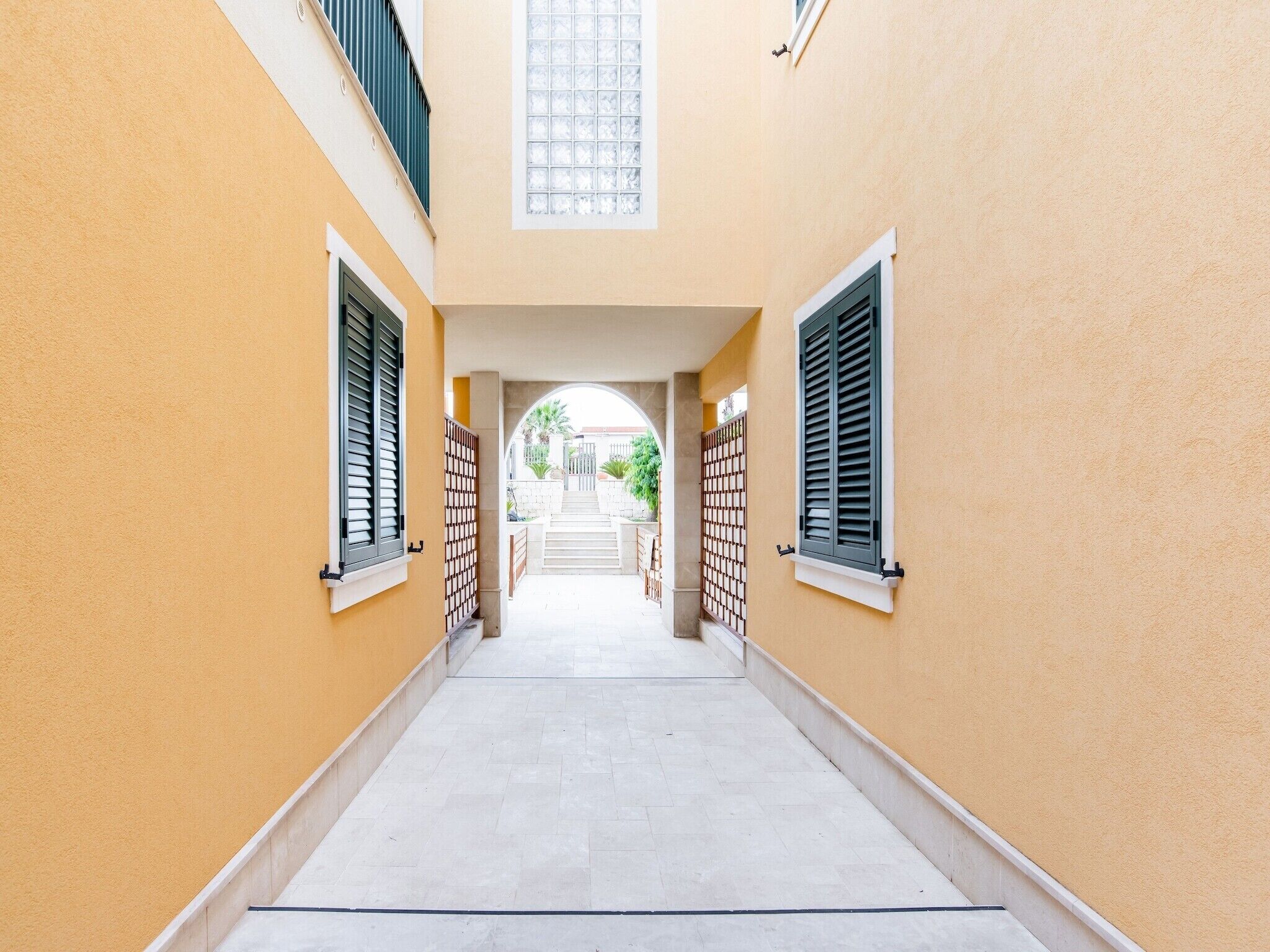 Ein schmaler, mit Fliesen ausgelegter Gang mit gelben Wänden und Fensterläden, der zu einer Treppe und einem Tor führt.