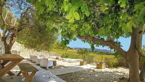 An outdoor seating area with a wooden table and chairs, surrounded by a stone wall and a view of a hilly landscape.