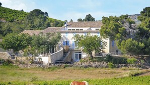 A two-story house with a stone wall and a tiled roof, surrounded by greenery and a field.