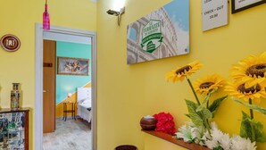 Ein Flur mit gelber Wand, einem gerahmten Bild, einem Regal mit Blumen und einem Schild, das die Frühstückszeiten anzeigt.